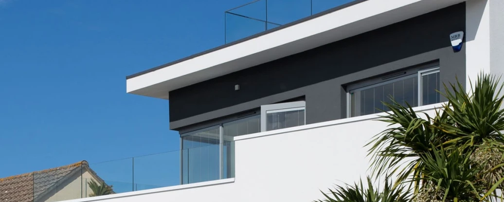 Modern two-story house with a flat roof and glass railing on the terrace. Clad in white and gray with large windows, surrounded by greenery under a clear blue sky.