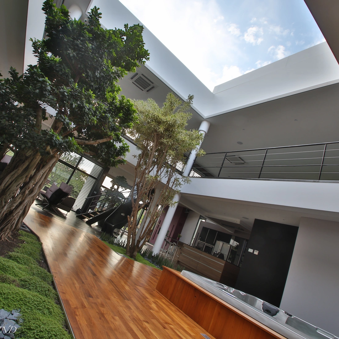 A modern interior space featuring an open layout with large windows, natural light streaming in, and indoor plants. A glossy wooden floor contrasts with green landscaping elements, while a sleek metal sculpture adds a contemporary touch.