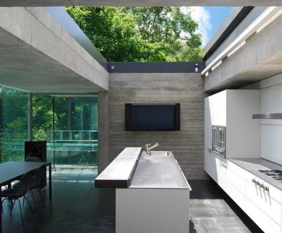 Cuisine moderne avec des murs en béton et des éléments en acier. Grandes fenêtres donnant sur un espace extérieur verdoyant. Un plan de travail en acier inoxydable et des appareils encastrés. Salle à manger adjacente avec des chaises et une table.