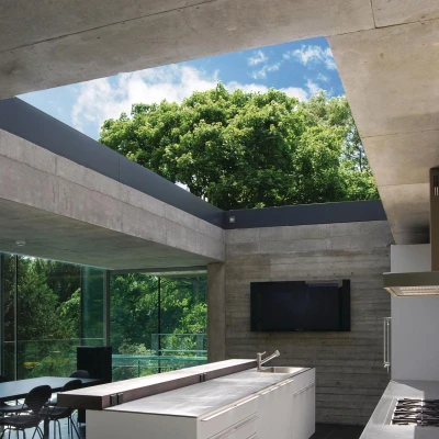 Cuisine moderne avec un grand plan de travail blanc, des armoires minimalistes et des fenêtres en verre laissant entrer la lumière naturelle. Un arbre verdoyant est visible à l’extérieur, créant une atmosphère harmonieuse avec l’espace.