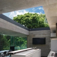 Cuisine moderne et épurée avec un grand îlot central, des murs en béton et des fenêtres offrant une vue sur la verdure extérieure. L’espace baigne dans la lumière naturelle.