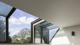 Two gray upholstered chairs are positioned in a well-lit room with large modern windows showcasing a blue sky and trees outside. Light wooden flooring complements the space, which has minimalist decor and built-in shelving.
