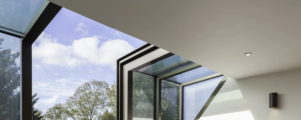 Deux fauteuils gris avec des pieds en bois sont placés dans une pièce lumineuse. De grandes fenêtres avec des toits en verre laissent entrer la lumière naturelle et offrent une vue sur des arbres à l’extérieur. Le sol est en bois clair.