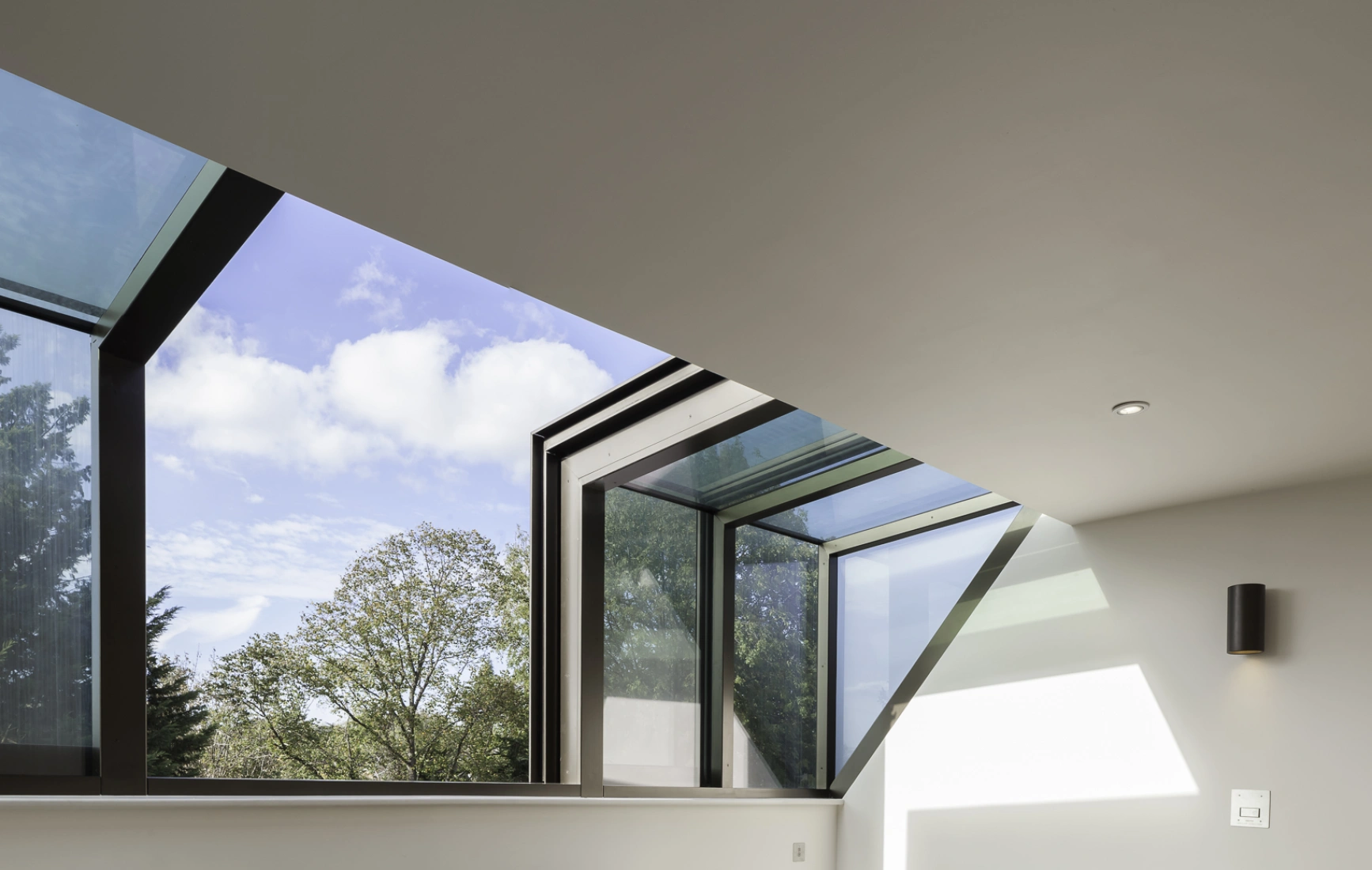 Espacio interior iluminado con dos sillones grises frente a una ventana grande con vidrio azul. El suelo es de madera clara y hay estanterías en la pared. Se pueden ver árboles y un cielo parcialmente nublado a través de la ventana.