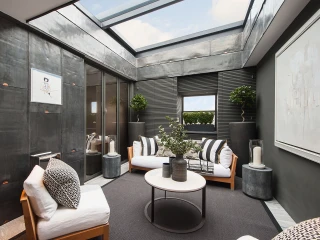 A modern indoor lounge area featuring minimalist furniture, including white sofas with black accent pillows, a round coffee table, and potted plants. Large windows and a skylight provide natural light. The walls are sleek and dark, with artistic decor enhancing the contemporary feel.