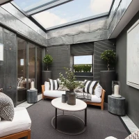 A modern sunroom featuring a sleek design with large skylights. Two sofas with white cushions and black decorative pillows are arranged around a round coffee table. The walls are a blend of dark materials, and there are potted plants and decorative elements enhancing the space. Natural light fills the room, creating a warm ambiance.