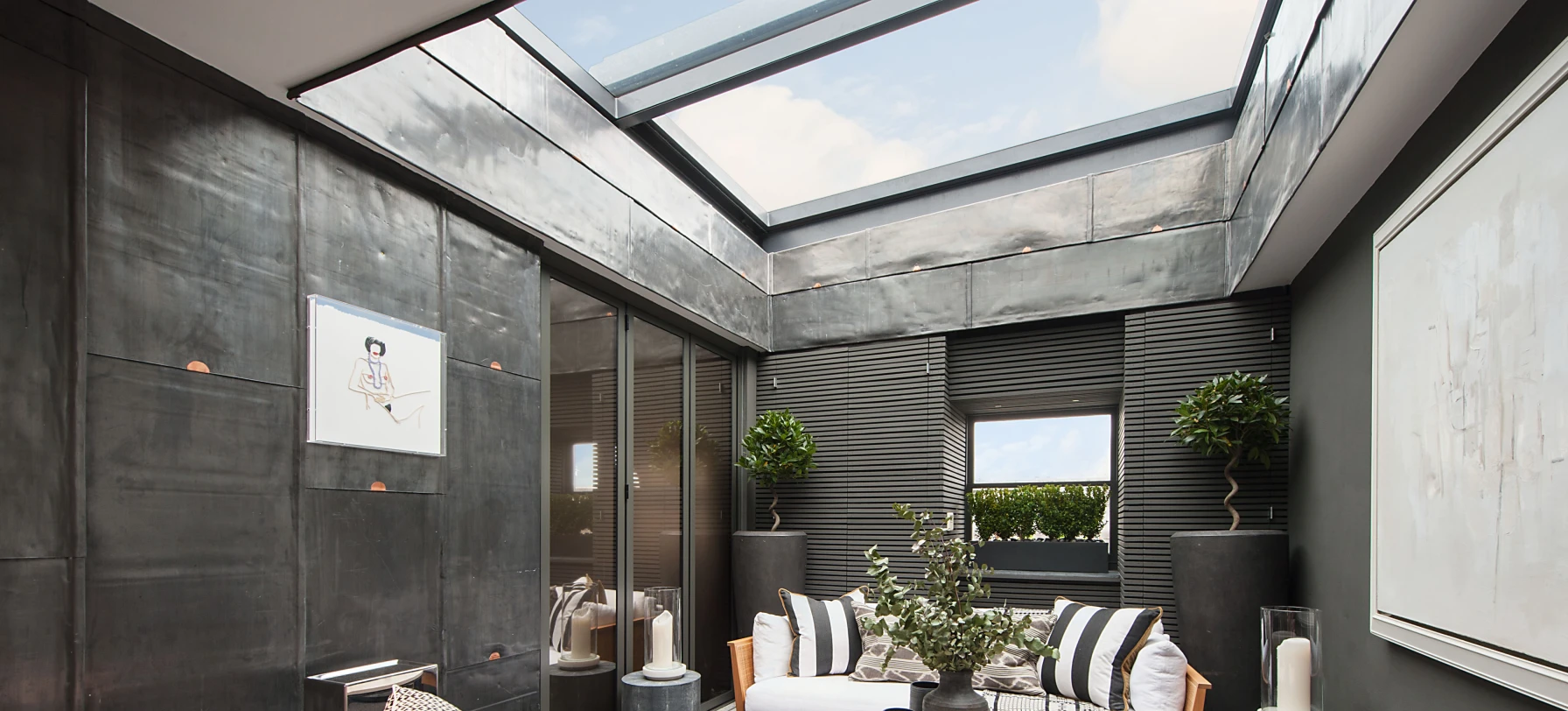 Espacio interior moderno con paredes de metal oscuro, sofá blanco y cojines decorativos. Hay una mesa de café circular y plantas en macetas. Una gran claraboya proporciona luz natural. Este ambiente combina elegancia y confort.