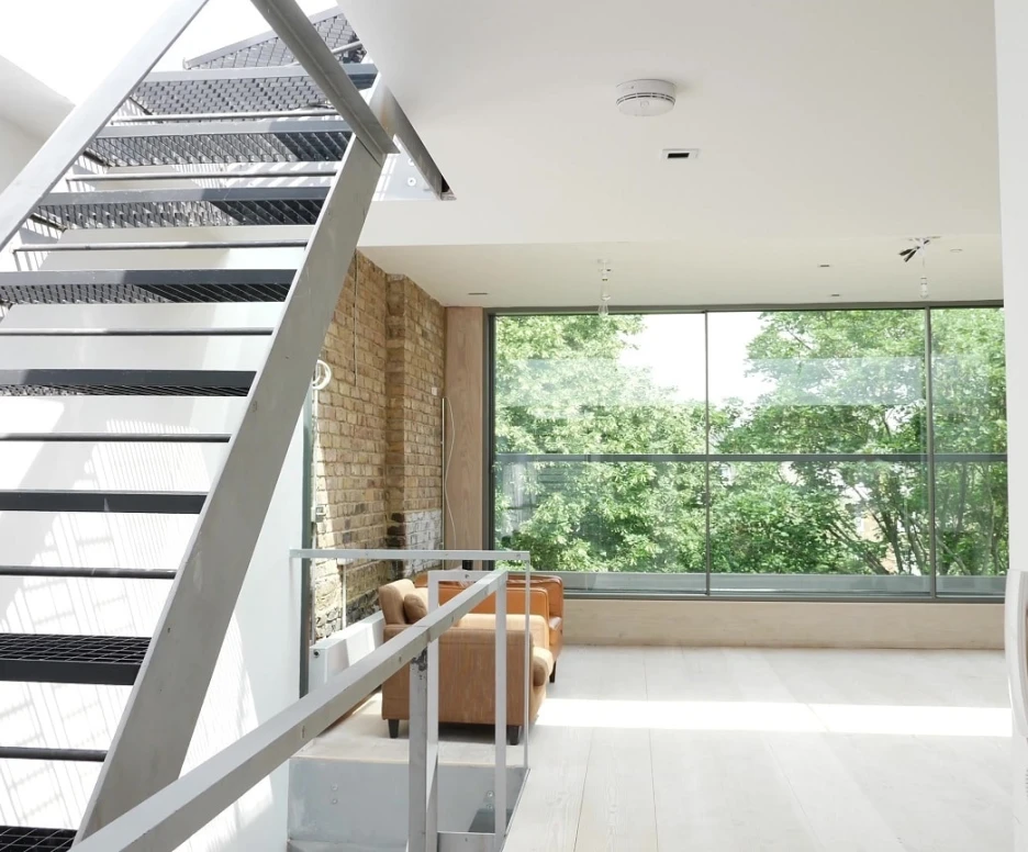 A modern staircase with a metal framework leads up to a bright, airy living space featuring large windows that overlook greenery. The interior has a combination of exposed brick and sleek white walls, with a cozy seating area visible in the background.