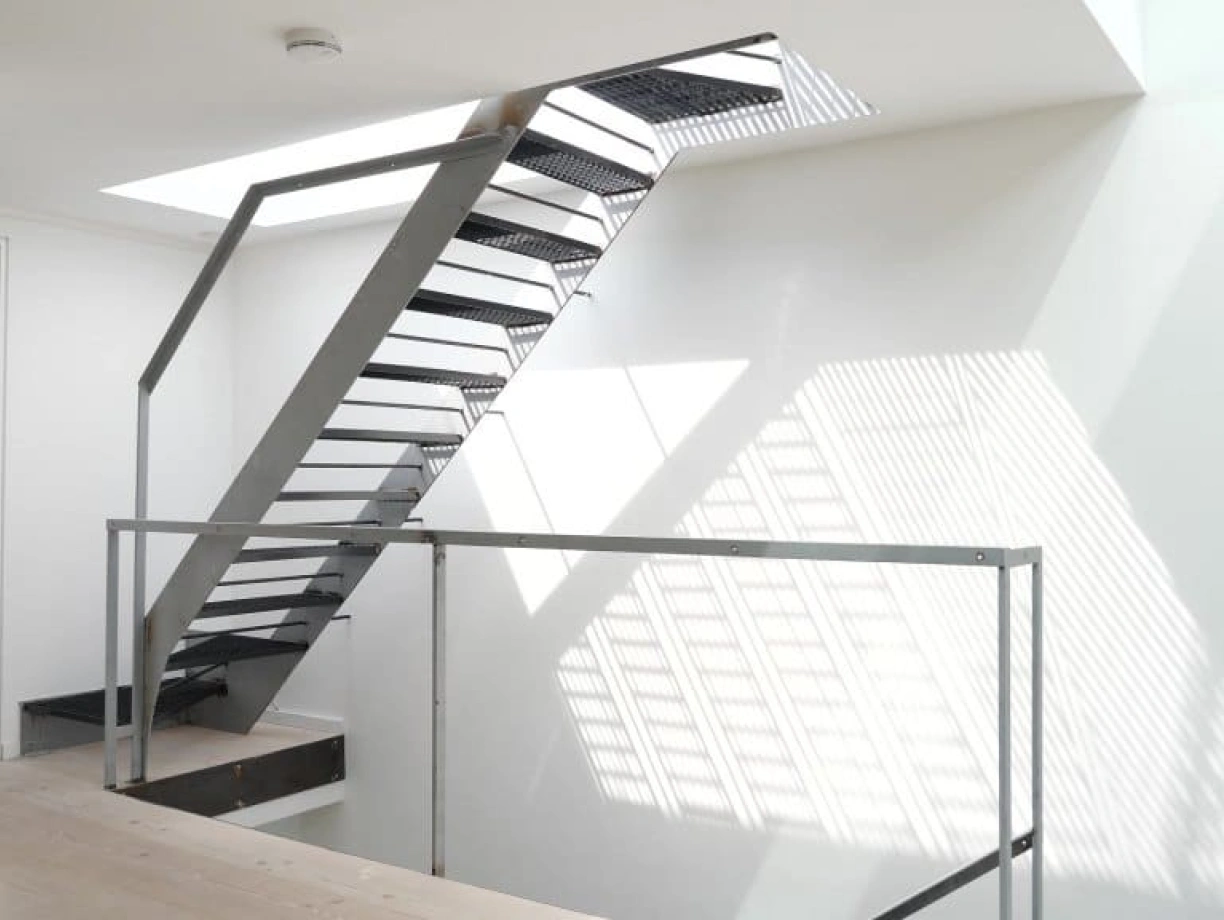 A modern staircase with a sleek metal frame and open treads, leading to a bright upper level. Natural light filters through a skylight, creating contrasting shadows on the white walls. A glass railing surrounds the staircase area.
