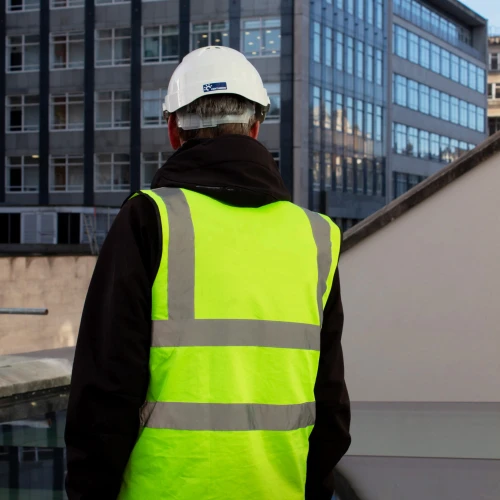 Hombre de espaldas, vistiendo un chaleco reflectante amarillo y casco blanco, observando un edificio moderno de oficinas.