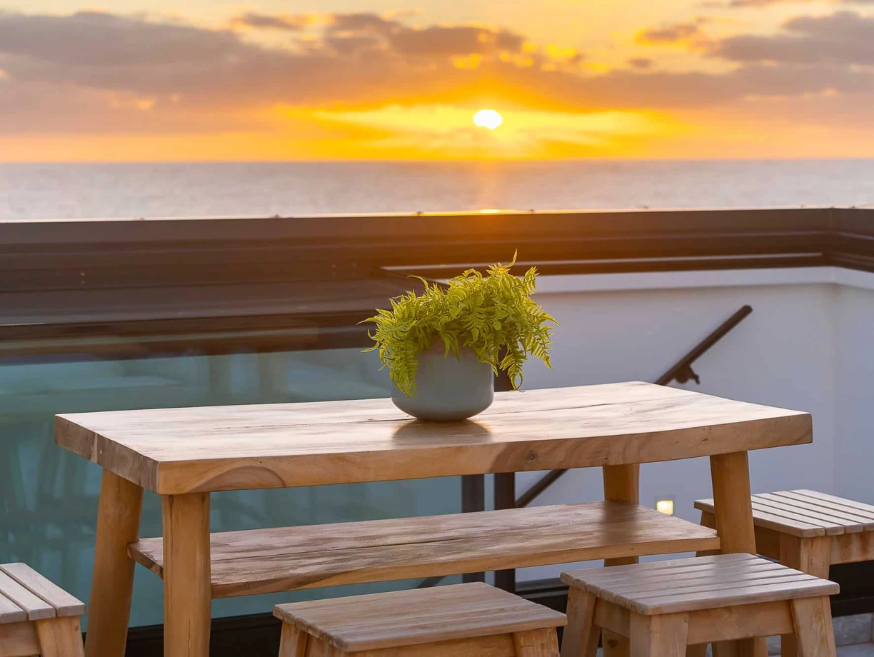 Un coucher de soleil sur l’océan, vu depuis un balcon avec une table en bois et des bancs. Un pot de plante verte est placé au centre de la table.