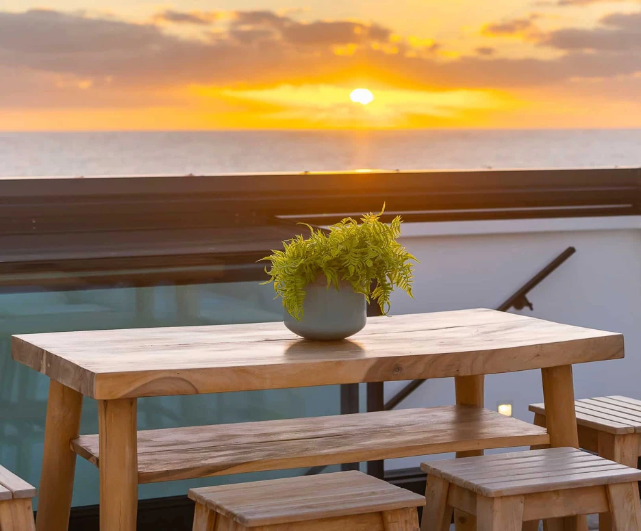 A wooden table with a potted fern sits on a rooftop terrace, overlooking a sunset over the ocean. The sky is painted with warm hues of orange and yellow.