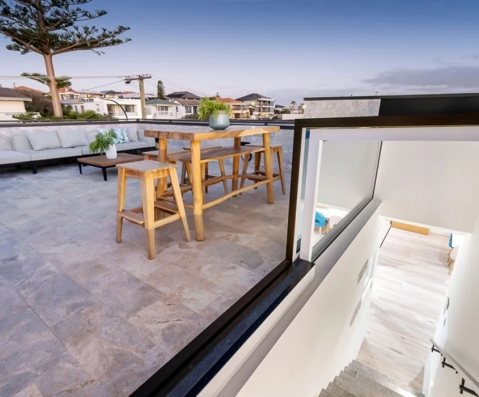 A rooftop terrace features a wooden dining table surrounded by chairs, with a comfortable lounge area and potted plants. In the background, a scenic view of rooftops and homes under a twilight sky. A staircase leads down into the interior.