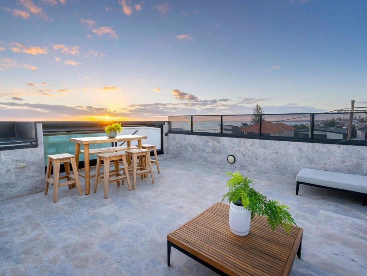 A rooftop terrace features a wooden dining table and chairs, a low wooden coffee table, and a potted plant. The scene is set against a vibrant sunset, illuminating the sky with shades of orange and pink. Surrounding buildings and a swimming pool are visible in the background.