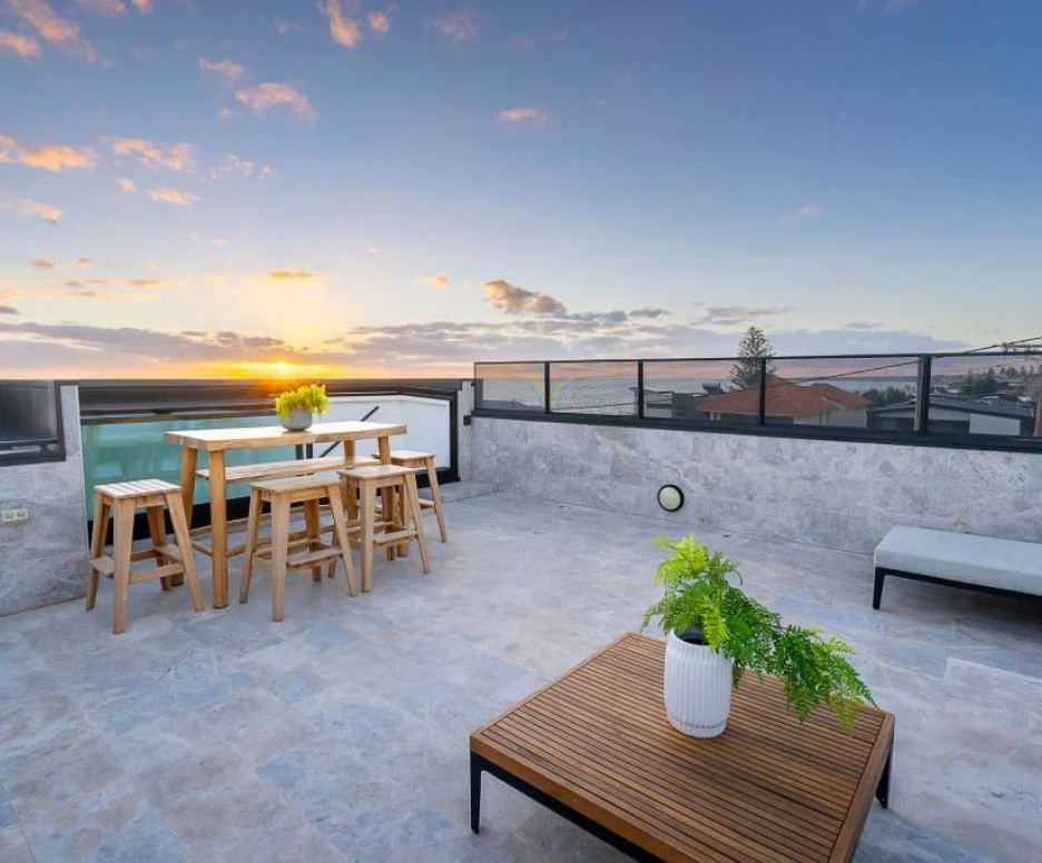 Terrasse moderne avec des tables en bois, un canapé et des plantes, surplombant un paysage urbain au coucher du soleil.