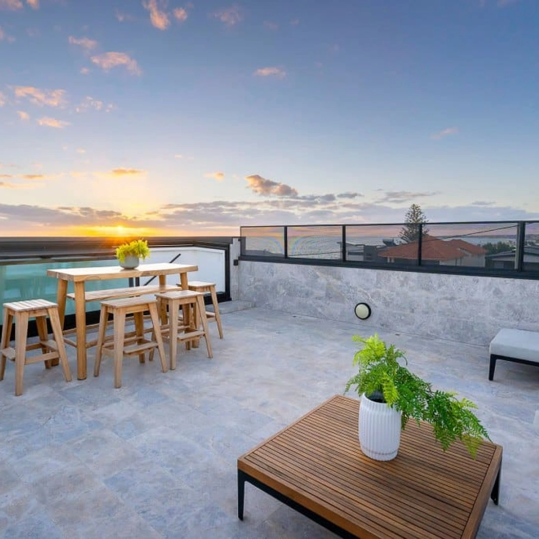 Terrasse moderne avec des tables en bois, un canapé et des plantes, surplombant un paysage urbain au coucher du soleil.