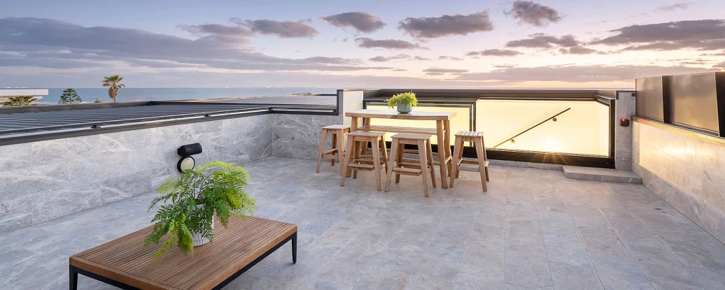 A modern rooftop terrace features a wooden table and stools surrounded by gray stone flooring. Lush green plants add a touch of nature, and the scene is set against a beautiful sunset sky with scattered clouds and a view of the ocean.