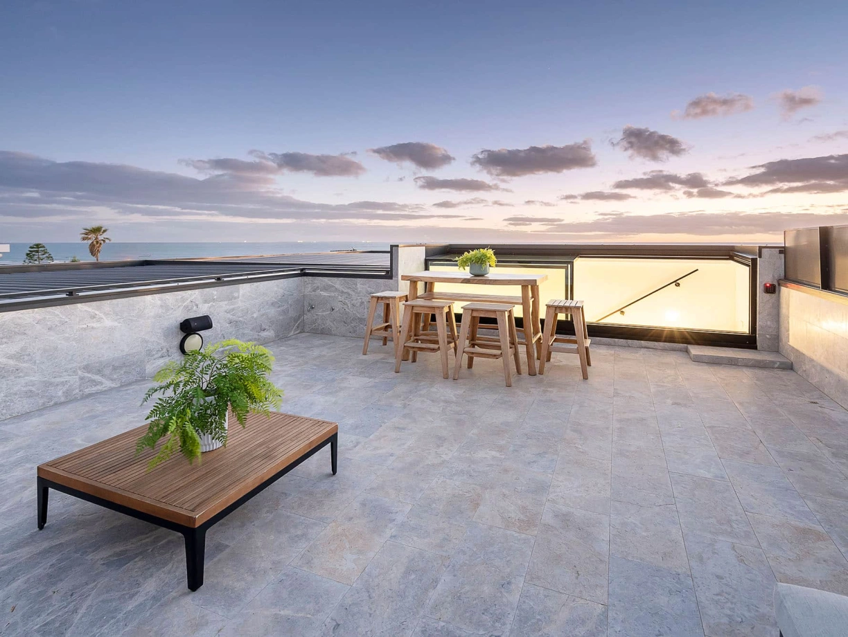 Terrasse spacieuse avec mobilier en bois, comprenant une table et des chaises, décorée de plantes. Le coucher de soleil se reflète sur l’horizon, créant une ambiance chaleureuse.