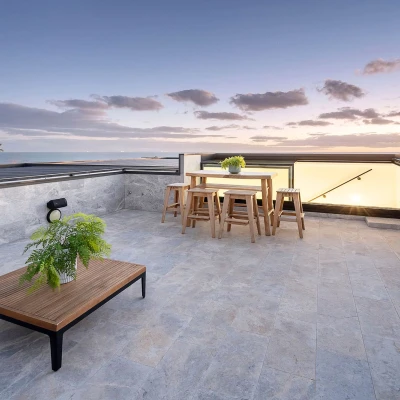 Terraza moderna con mesas y sillas de madera, decorada con plantas, y vistas al océano bajo un cielo colorido al atardecer.