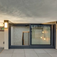 A modern rooftop entrance with large glass doors revealing a staircase. The sky is overcast with hints of sunset colors in the background. Stylish outdoor lighting fixtures flank the entrance.