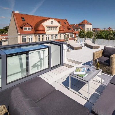 Terraza moderna con muebles de exterior cómodos, incluyendo un sofá y una mesa, con una vista clara del cielo y edificios cercanos. Se aprecian áreas ajardinadas y una piscina al fondo.
