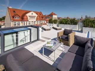A stylish rooftop terrace features modern outdoor furniture, including a cozy sectional sofa and a small table. The space is surrounded by greenery, with a city skyline and a clear blue sky in the background.
