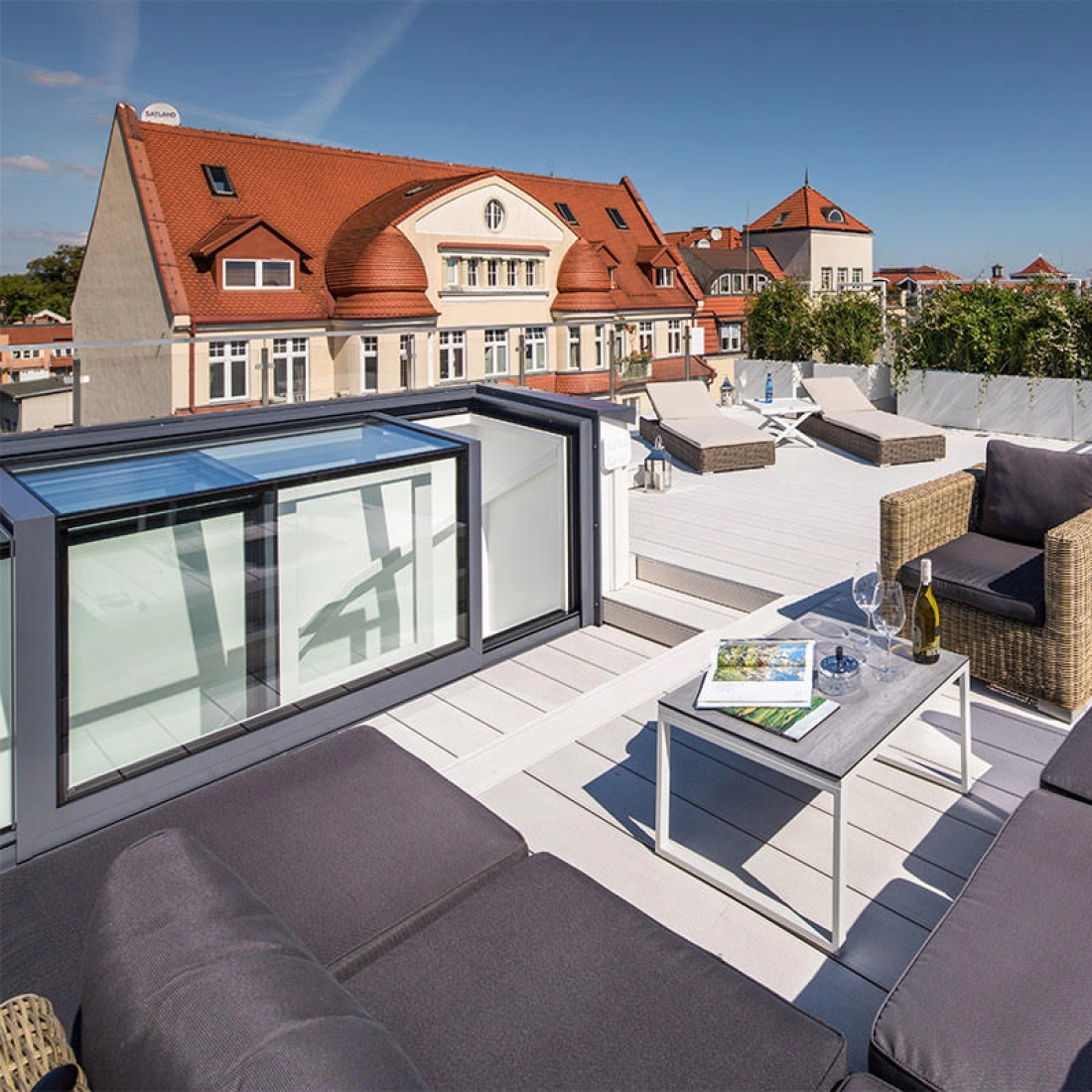 A stylish rooftop terrace features modern gray seating, a small table with a magazine and two glasses, surrounded by greenery and a clear blue sky. In the background, a historic building with a red roof is visible.