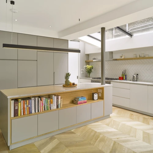 Moderne, hell erleuchtete Küche mit hölzernem Boden, einer Kücheninsel mit offenen Regalen für Kochbücher und einer schlichten, eleganten Farbkombination aus Grau und Weiß. Große Fenster sorgen für viel Tageslicht.