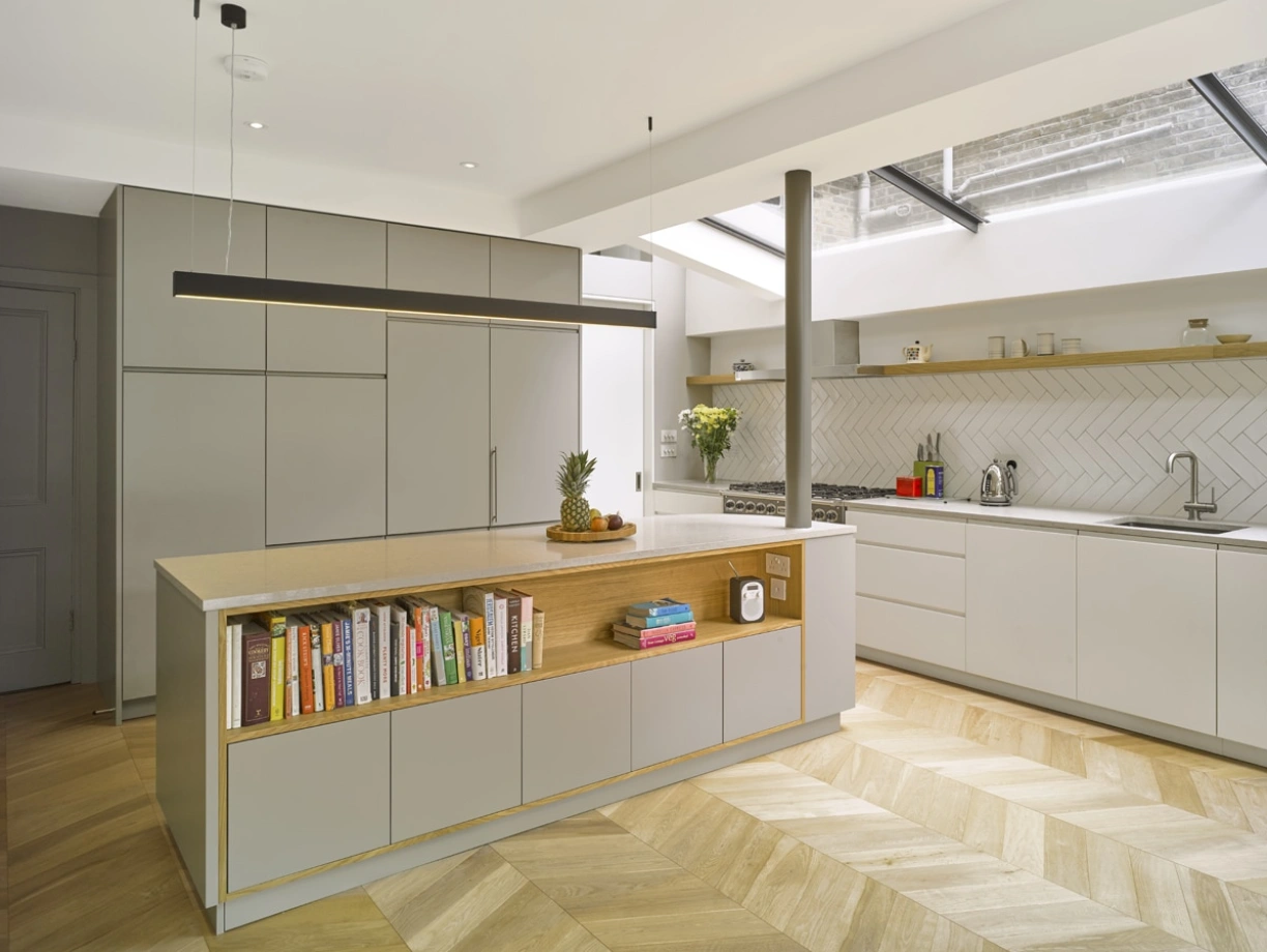 Cuisine moderne avec un îlot central en bois et un espace de rangement. Étagères ouvertes avec des livres de cuisine. Éclairage naturel provenant d’une fenêtre de toit. Sol en bois à motifs chevron.