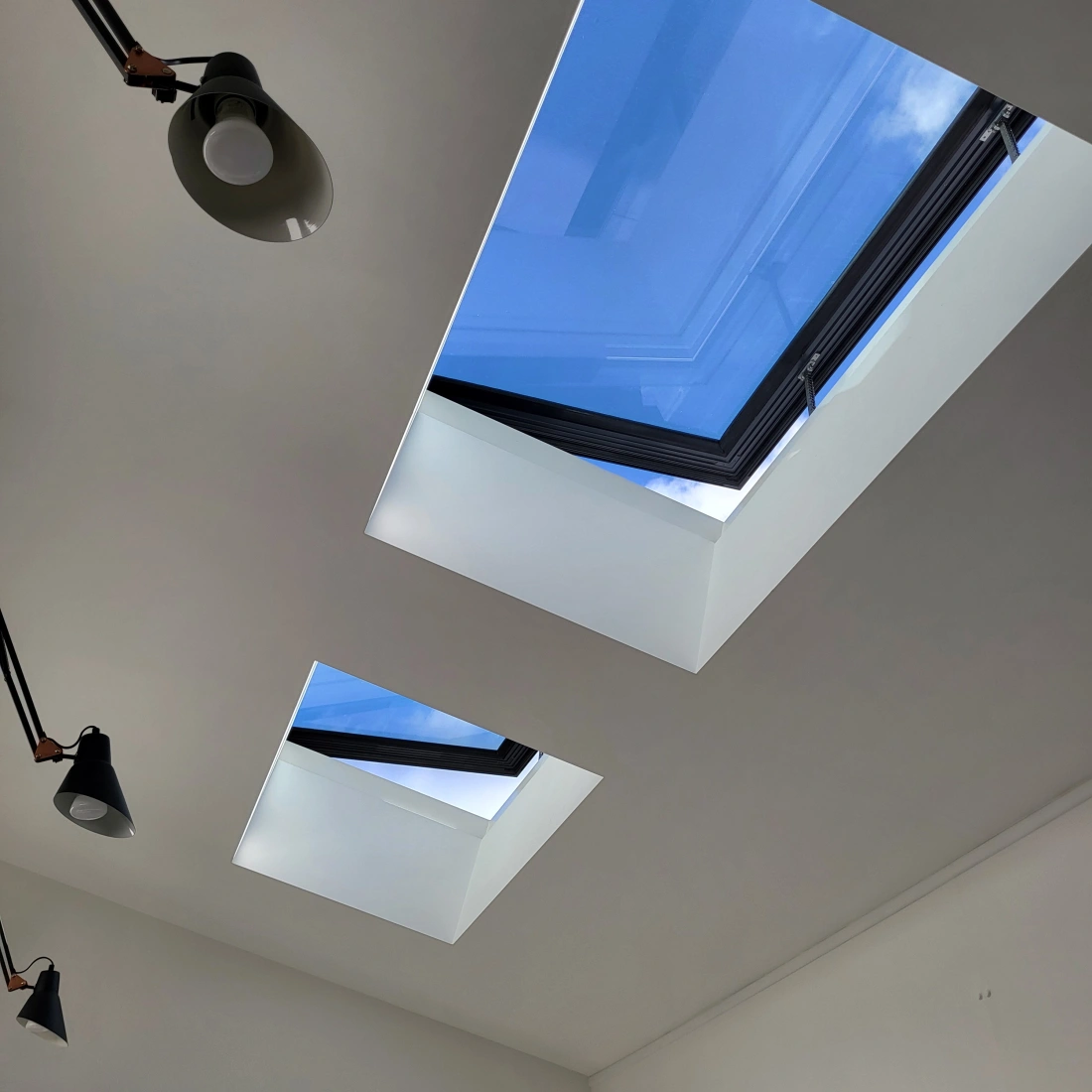 Two skylights in a modern room ceiling, framed by black borders, letting in natural light and revealing a clear blue sky. Adjustable pendant lights are mounted on the ceiling.