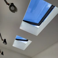 Two skylights in a modern ceiling allow natural light to enter, revealing a clear blue sky and few clouds. The ceiling is smooth and white, with sleek lighting fixtures along the sides.