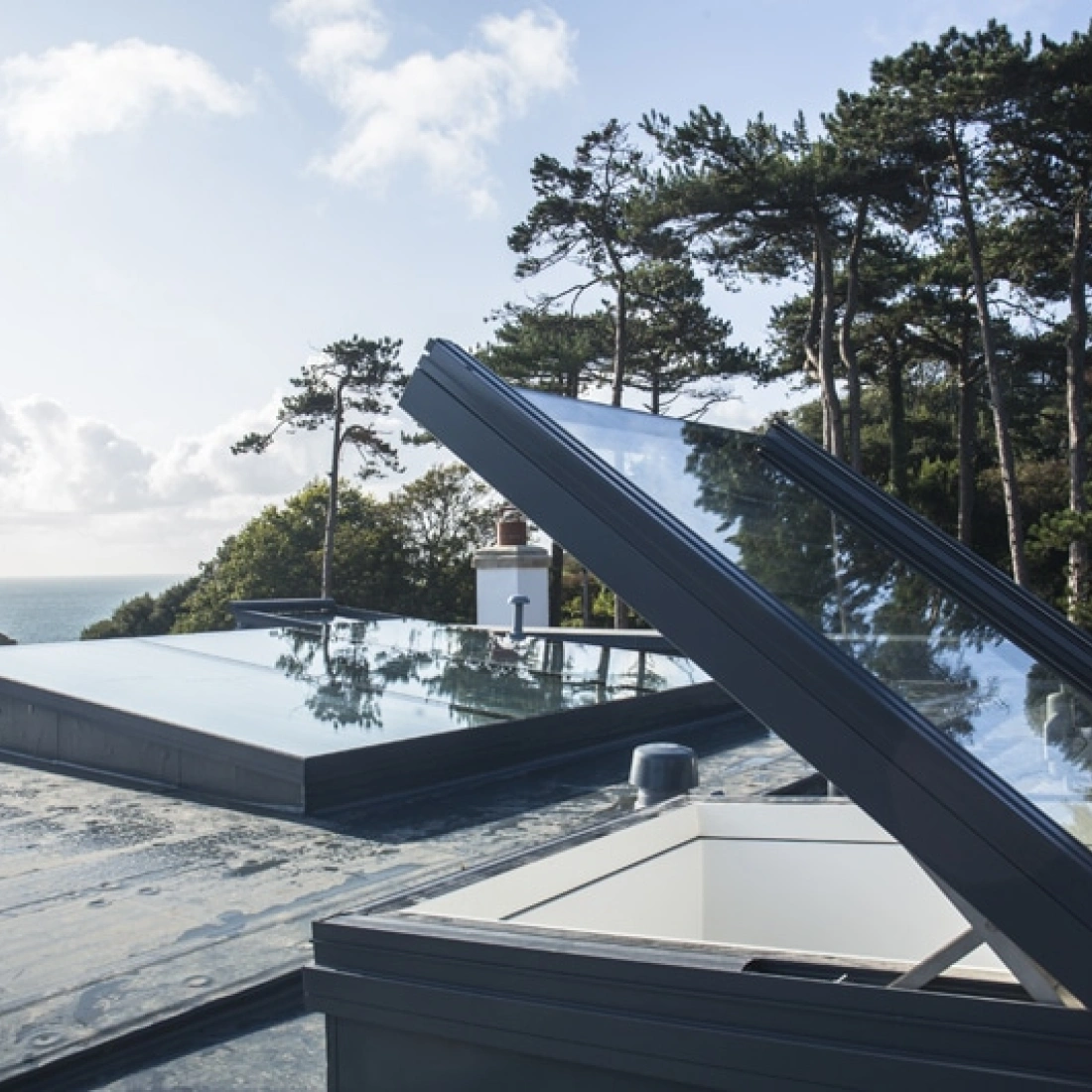 Des fenêtres de toit ouvertes sur une terrasse, avec une vue panoramique sur la mer et des arbres en arrière-plan sous un ciel ensoleillé.