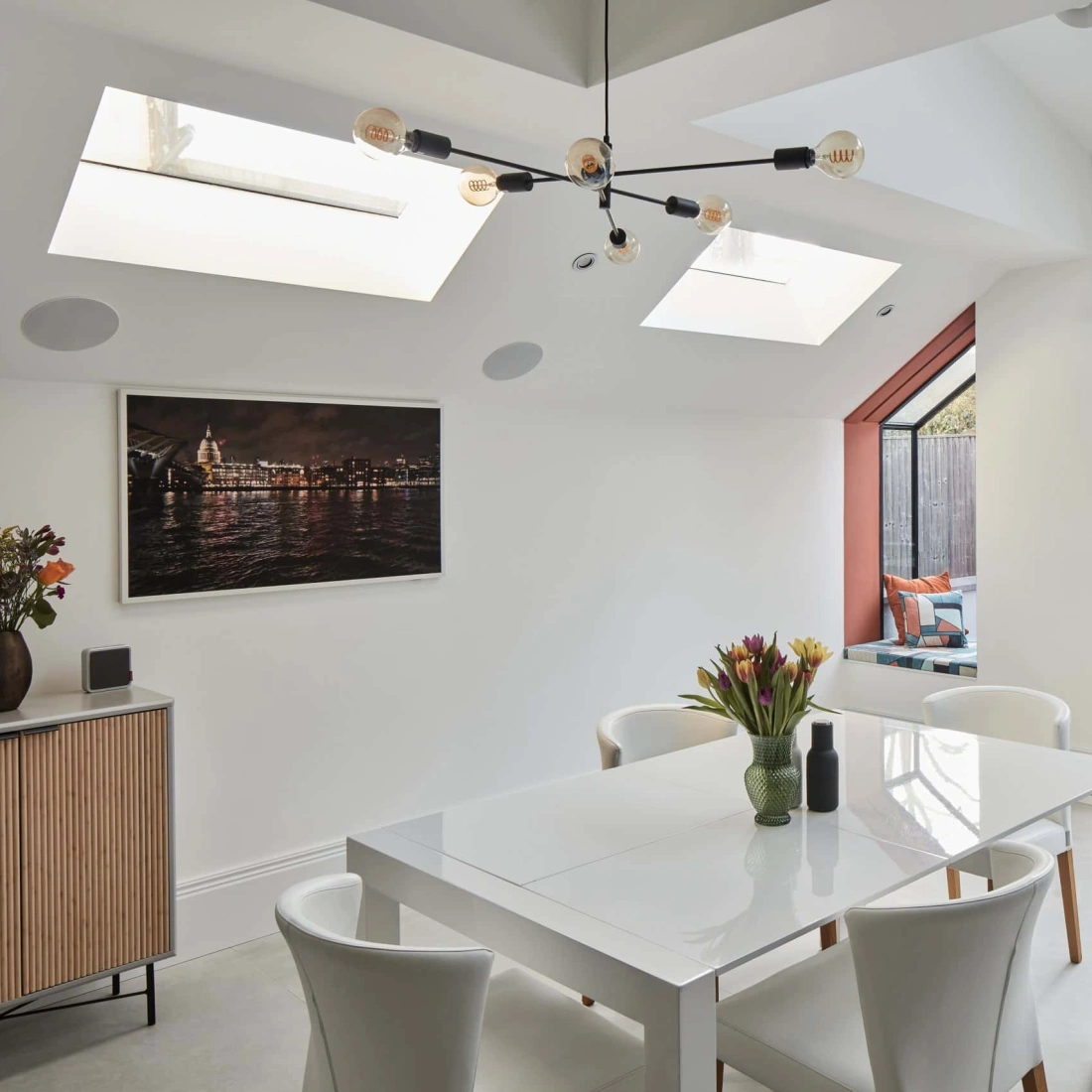 Bright and modern dining area featuring a white table surrounded by four chairs, a floral centerpiece, and large skylights. A sideboard with wood detailing and a photograph adorn the wall, while a glass door opens to a backyard with greenery.