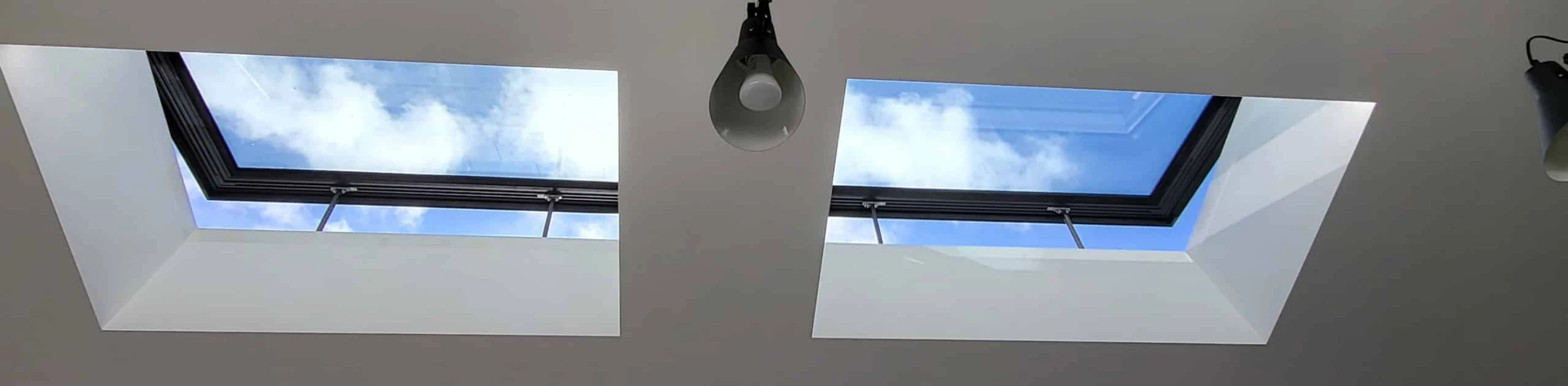 Dos tragaluces en un techo blanco permiten la entrada de luz natural. El cielo azul y algunas nubes son visibles a través de los tragaluces. Hay lámparas colgando en el techo.