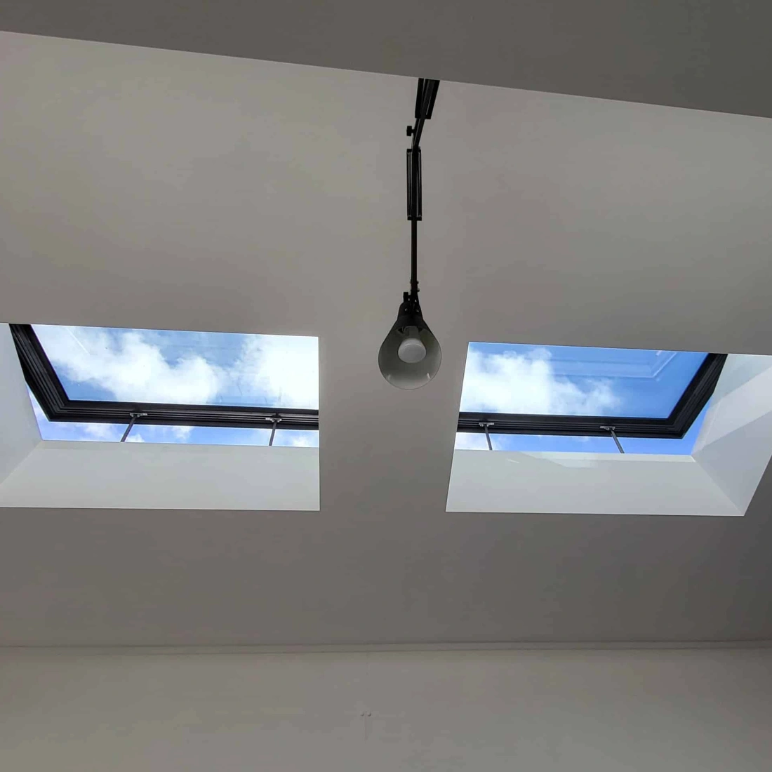 Two square skylights in a white ceiling, revealing a bright blue sky with clouds above. A light fixture hangs below one of the skylights.