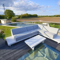 Moderne Terrasse mit einer grauen Sitzgarnitur, einem weißen Tisch und einer Glasunterlage. Im Hintergrund sind Wiesen und Bäume zu sehen, unter einem blauen Himmel mit Wolken.