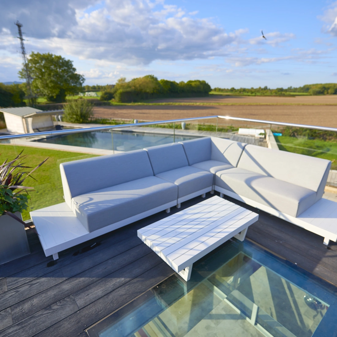 A modern outdoor seating area featuring a gray sectional sofa and a white coffee table on a deck. The background shows a lush landscape with fields, trees, and distant buildings under a partly cloudy sky.