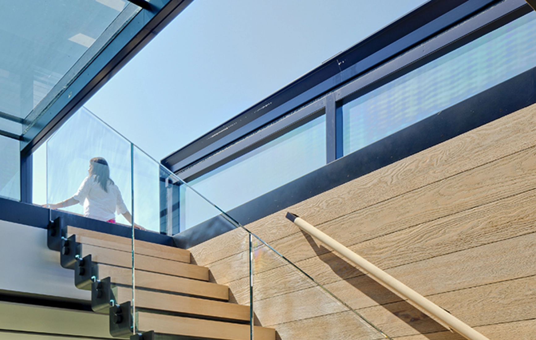 A modern staircase with wooden steps and glass railing, leading to a bright upper floor. Natural light floods the space, highlighting the contemporary design.