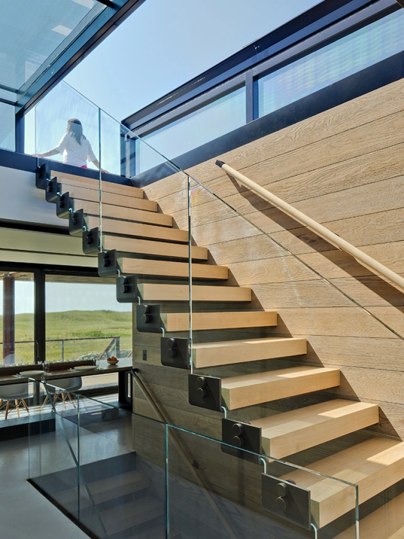Escalera moderna con peldaños de madera y barandilla de vidrio. Al fondo, una persona se asoma por un ventanal que deja entrar luz natural. El ambiente es minimalista y contemporáneo.