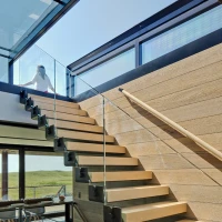A modern staircase with wooden steps and a glass railing leads up to a bright, open space with large windows. A person is seen ascending the stairs, with sunlight streaming in from above.