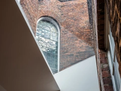 A view of a brick wall with a large arched stained glass window. Light streams through the window, illuminating the space. A diagonal white ceiling contrasts with the rustic brick.