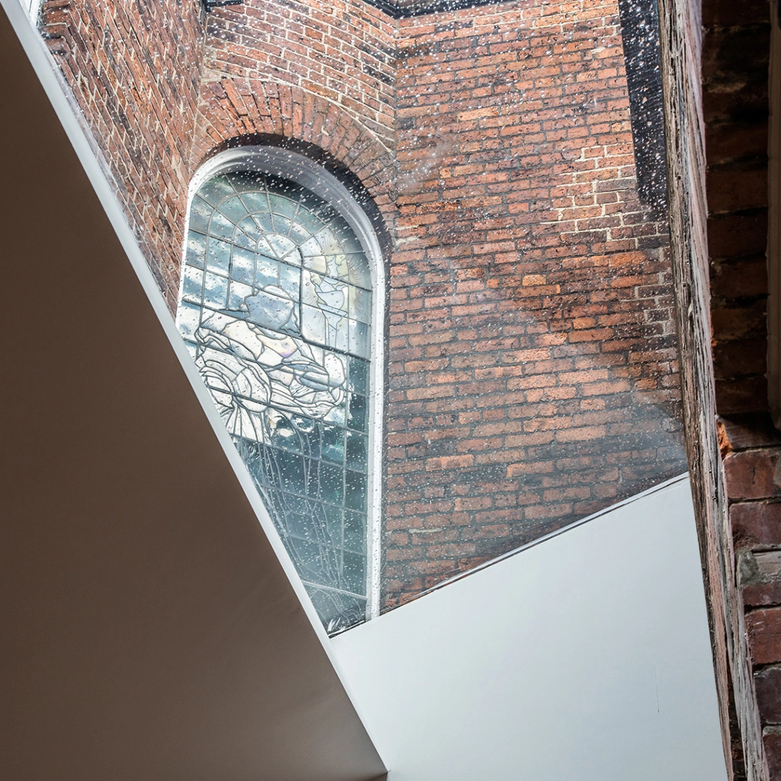 A view of a brick wall with a large arched stained glass window. Light streams through the window, illuminating the space. A diagonal white ceiling contrasts with the rustic brick.
