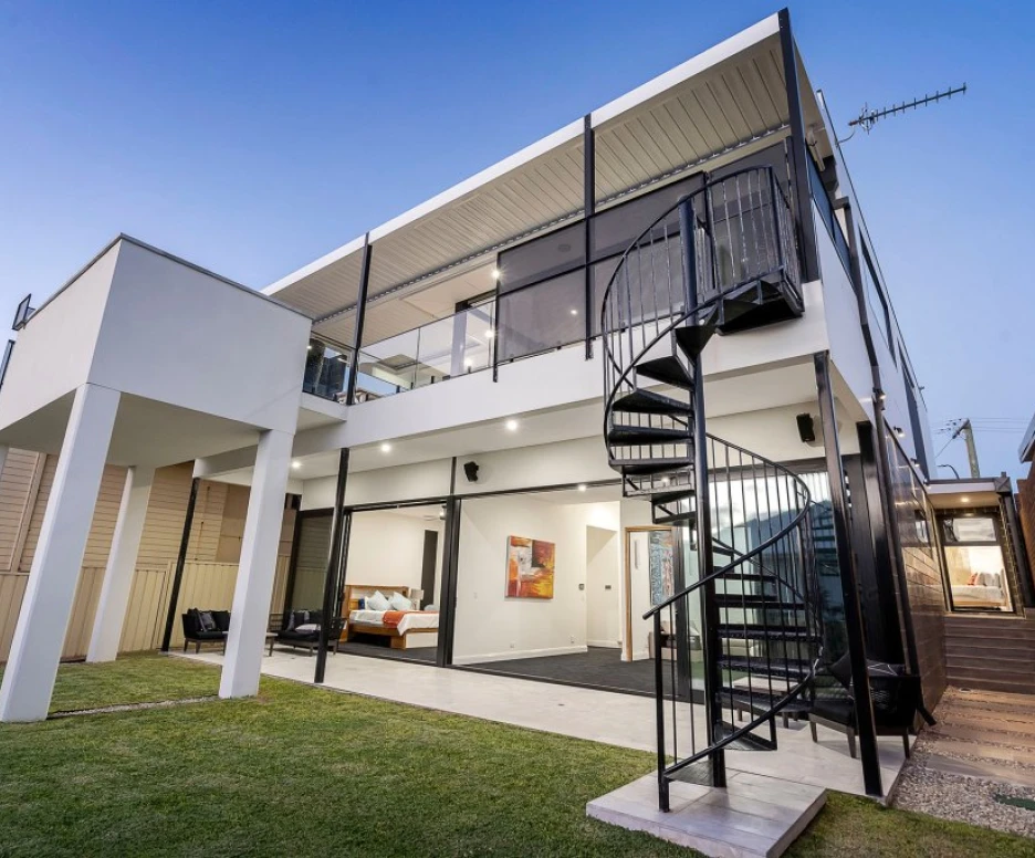 A modern two-story house with a spiral staircase, large glass windows, and an outdoor lawn area. The exterior features a sleek, minimalist design with a combination of white and black elements.