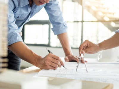 Dos personas están trabajando juntas en un plano arquitectónico. Una persona utiliza un compás para medir mientras la otra dibuja con un lápiz. En la mesa hay herramientas de diseño y un casco de construcción. La luz natural entra por las ventanas, creando un ambiente luminoso.