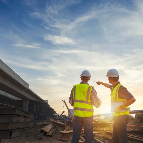 Two construction workers in safety vests and helmets observe a construction site at sunset, discussing plans while pointing towards the horizon.