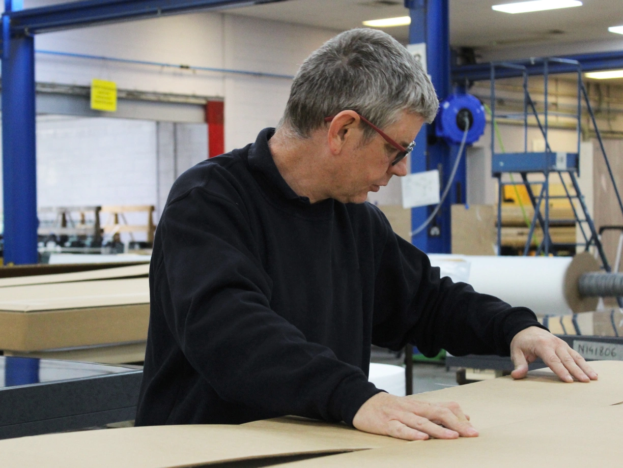 A person in a black sweater is carefully inspecting large sheets of cardboard in a warehouse environment. The background shows industrial equipment and shelving.