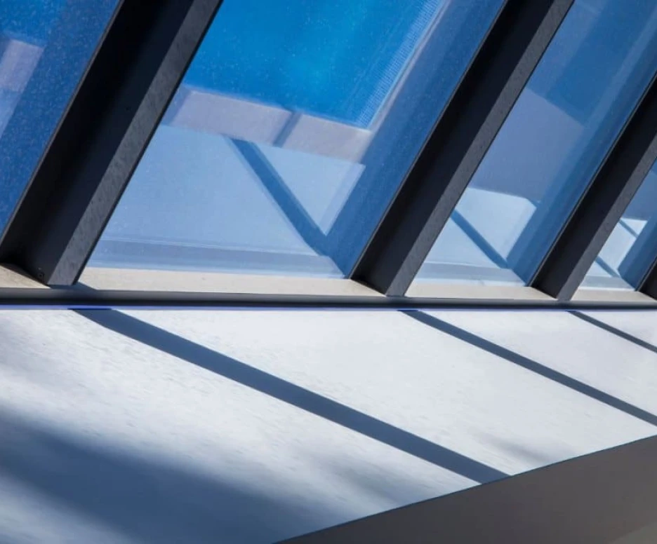 Sunlight streams through large, angled glass panels, casting geometric shadows on a white surface below. The clear blue sky is visible in the background.