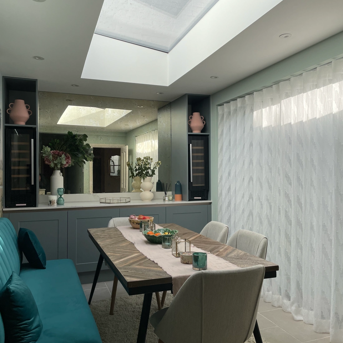 A modern dining area featuring a large table with a light-colored runner, surrounded by stylish chairs. The room has a skylight letting in natural light, and a soft rug covers the floor. Decorative plants and vases adorn the space, contributing to a bright and inviting atmosphere.