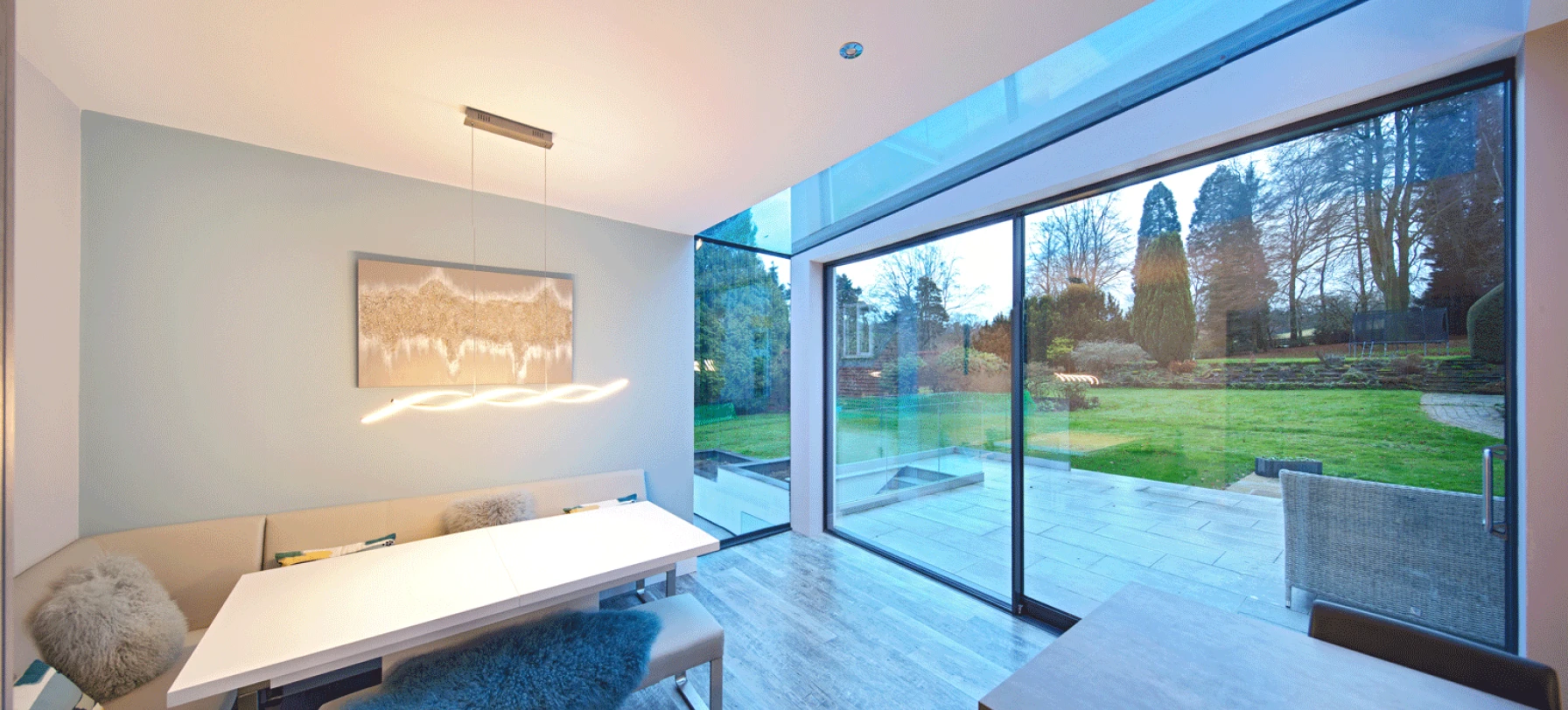 A modern dining area featuring a sleek white table and soft seating, with a large glass wall providing a view of a lush garden outside. Soft lighting hangs above the table, creating a warm atmosphere.