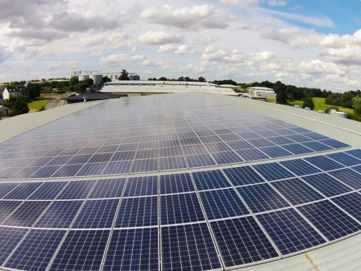 A wide view of a rooftop covered in solar panels, reflecting sunlight under a partly cloudy sky. In the background, residential buildings and green fields are visible.